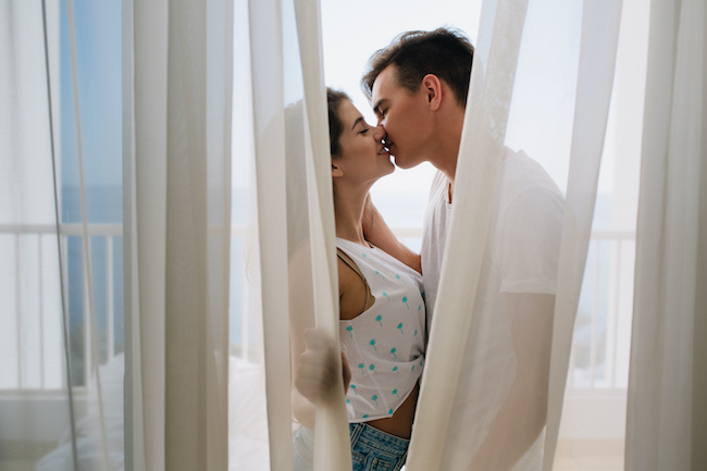 Graceful girl in white tank-top gently kissing her brunette boyfriend hiding behind light curtains. Portait of romantic young couple spending time together on balcony enjoying each other..